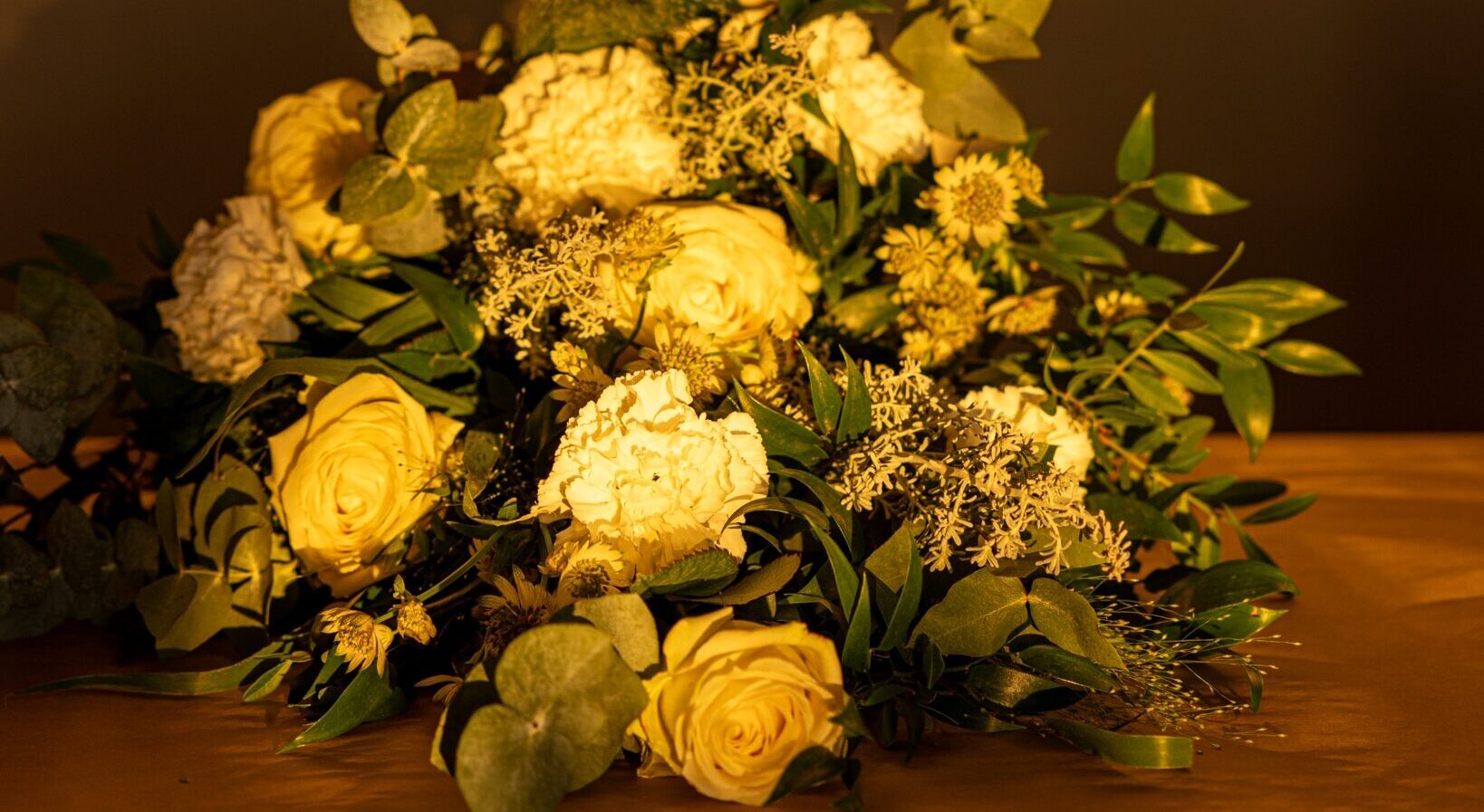 Kaunis surulaite, joka on asetettu arkun kannen päälle. Aurinko paistaa lämpimästi ja heijastaa valoa valkoisiin ruusuihin kukkalaitteessa, luoden arvokkaan tunnelman.