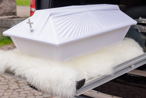 Kuva lapsen valkoisesta arkusta vainajan kuljetusautossa, asetettuna kauniin valkoisen taljan päälle. Arkku luo kunnioittavan ja rauhoittavan tunnelman.
